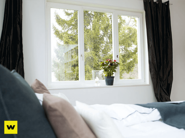 Helles Schlafzimmer mit großem Fenster, Vorhängen und grüner Zimmerpflanze auf der Fensterbank.