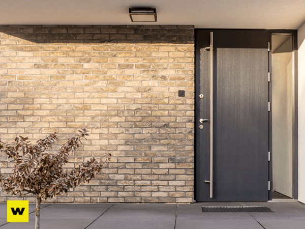 Moderne Haustür aus Aluminium neben heller Klinkerfassade und kleiner Pflanze im Eingang.